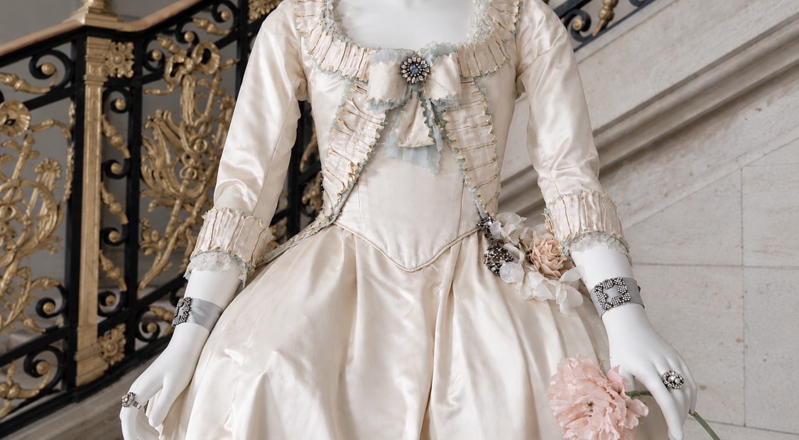 Detail of an ivory silk court gown from Sofia Coppola’s Marie Antoinette, trimmed with pleated ruffles, a jeweled brooch and silk flowers